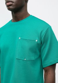 Τιρκουάζ βαμβακερό t-shirt με κοντά μανίκια και ραμμένο τσέπη που διαθέτει ασημένια κλείσιμο snap στο αριστερό στήθος. Απαλή υφή, χαλαρή εφαρμογή.