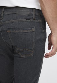 Mörkgrå jeans med orange sömmar, synlig bakficka med en dekorativ söm och en svart etikett på midjan. Nära bild av sidovyn.