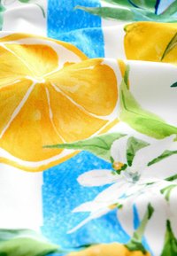 Heller Stoff mit gelben Zitronenschnitten, grünen Blättern, weißen Blumen und blauen Streifen, der Falten und Texturdetails zeigt.