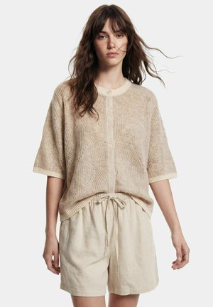 Kvinde med langt brunt hår iført en beige kortærmet knaptrøje og lysebeige shorts med snøre foran en ensfarvet baggrund.