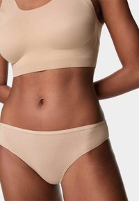 Mujer vistiendo un sujetador beige sin costuras y ropa interior a juego, de pie con las manos detrás de la espalda contra un fondo liso.