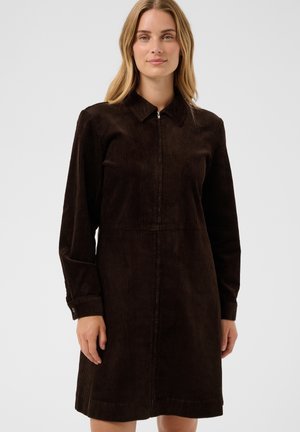 Femme portant une robe longue à manches longues en velours côtelé marron foncé avec une fermeture éclair à l’avant et un col, debout devant un fond clair uni.