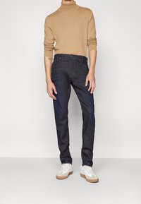 Jeans in denim blu scuro con vestibilità slim, abbinati a un maglione a collo alto beige chiaro. Le sneakers presentano dettagli in pelle bianca e scamosciato beige.