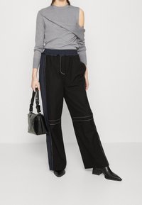 Haut gris à manches longues avec un design asymétrique et une épaule dénudée ; associé à un pantalon noir ample avec des coutures blanches et une taille cintrée.