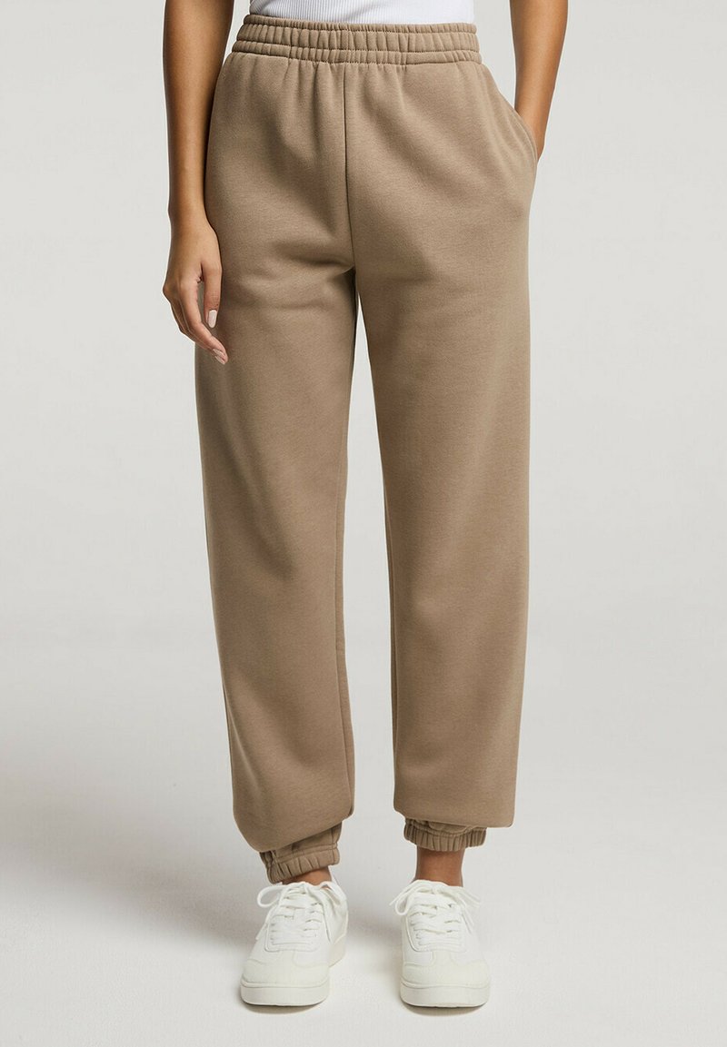 Beżowe dresy z elastycznym pasem, kieszeniami bocznymi i ściągaczami przy kostkach, w połączeniu z białymi sneakersami. Miękki materiał i luźny krój.