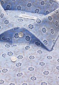 Chemise bleu clair avec un petit motif floral bleu et blanc, boutons blancs et une étiquette visible de la marque "seidensticker" à l'intérieur du col.