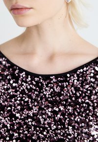 Top s flitrami v purpurovej a striebornej farbe s hladkou textúrou, orámovaný čiernym výstrihom. Detailný záber na látku.