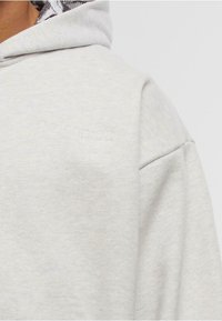 Sudadera gris claro con el sutil logo "COLUCCI" bordado en el pecho, mostrando el hombro, parte del cuello y la parte superior del brazo.