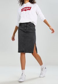 Falda lápiz de mezclilla negra con una abertura frontal, combinada con una sudadera blanca y zapatillas deportivas. Presenta un diseño ajustado y detalles de costura visibles.