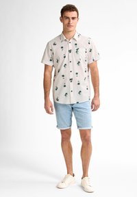 Camicetta a maniche corte color crema con stampa di palme verdi e fiori, abbinata a pantaloni di jeans leggeri e scarpe da ginnastica color avorio.