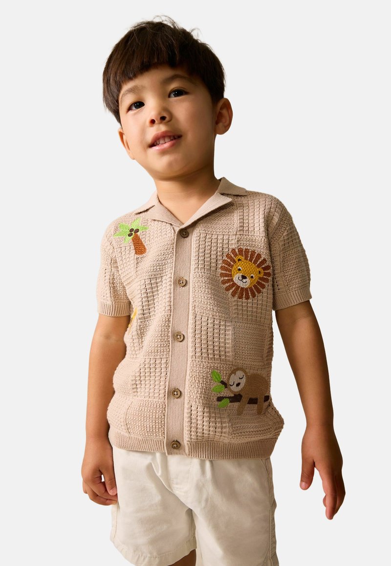 Camiseta de punto beige claro de manga corta con un patrón texturizado, que presenta bordados de león, árbol y perezoso. Tres botones en la parte delantera.