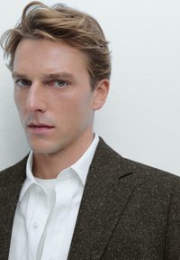 Mann mit kurzem, hellbraunem Haar, der ein dunkel strukturiertes Blazer über einem weißen Hemd mit Knöpfen trägt, vor einem schlichten weißen Hintergrund.