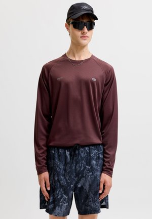 Junger Mann trägt ein bordeauxrotes Langarmshirt, dunkle gemusterte Shorts, schwarze Kappe und schwarze getönte Sonnenbrille, steht vor einem einfachen Hintergrund.