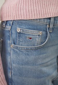 Vaqueros de mezclilla azul claro con un bolsillo trasero visible, con un pequeño logo en rojo, blanco y azul. Combinados con un suéter rosa acanalado.