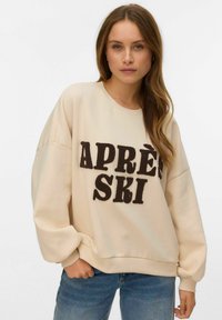 Helebeež värvi jakk, oversized lõikega, millel on suur tumepruun tekst "APRÈS SKI" tekstuurse kangaga, soonikkoes kaelus ja mansetid ning vabaaja stiil.