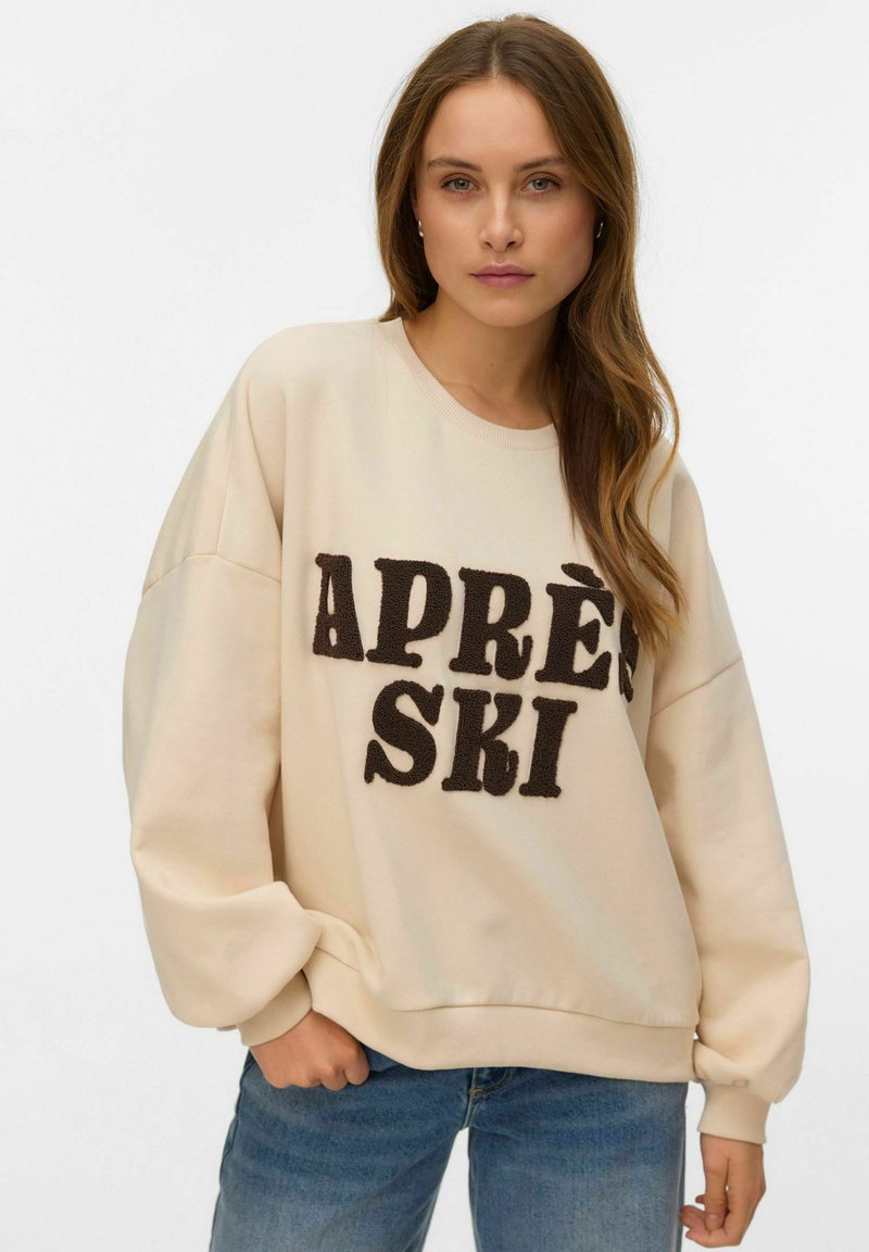 Helebeež värvi jakk, oversized lõikega, millel on suur tumepruun tekst "APRÈS SKI" tekstuurse kangaga, soonikkoes kaelus ja mansetid ning vabaaja stiil.