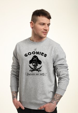 Man with short brown hair and ear piercings wearing gray sweatshirt with skull, crossed swords, and "The Goonies Never Say Die" text.
