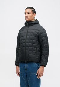 Black padded jacket featuring a hood, quilted design, zippered pockets, and a ribbed cuff. Worn over blue denim pants.