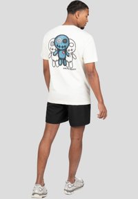 Camiseta blanca con un gráfico azul y negro de personajes de dibujos animados en la parte posterior. Mangas cortas, corte relajado, usada con pantalones cortos negros.