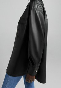 Black leather shirt with long sleeves, button closure, and patch pocket. Smooth texture and structured fit, paired with denim.