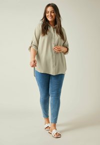 Hellgrünes Hemd mit hochgerollten Ärmeln, kombiniert mit eng anliegenden blauen Jeans und weißen Sandalen. Glattes Material, lockere Passform und klassisches Design.