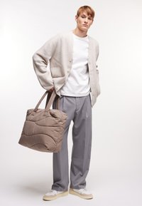 Młody mężczyzna w beżowym kardiganie, białej koszulce, szarych spodniach i białych trampkach, trzymający dużą taupe pikowaną torbę.