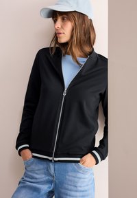 Svart zip-up jacka med randiga ärmar, i kombination med en ljusblå t-shirt och denimjeans. Accentuerad med en mjuk blå keps.
