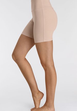 Beine und Hüften einer Person, die beige eng anliegende Shorts trägt, steht auf einem Fuß, während der andere Fuß auf den Zehenspitzen erhoben ist.