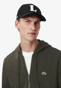 Casquette noire avec un logo "L" blanc, portée avec un sweat à capuche zippé vert avec un petit logo de crocodile vert sur la poitrine et une chemise blanche en dessous.