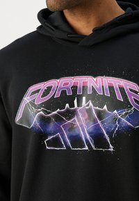 Melna kapuce ar lielu, krāsainu "FORTNITE" grafiku, kas rotāta ar purpursarkanām un ziliem akcentiem, zibens motīviem un galaxijas iedvesmotu fonu.