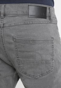 Pantaloni in denim grigio caratterizzati da una texture liscia, una tasca posteriore con cuciture sottili e un'etichetta in pelle nera sulla cintura.