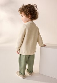 Cream ribbed knit sweater and light green trousers, paired with beige sneakers, child standing next to a white cube. Soft textures.