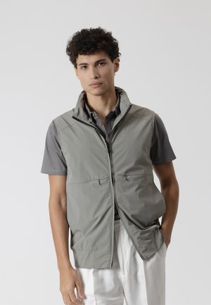 Joven con cabello rizado que lleva un chaleco gris con cremallera sobre una camiseta gris de manga corta y pantalones blancos sobre un fondo liso.