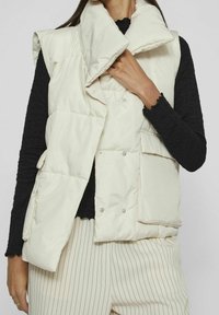 Lättviktskräm puffer väst med stor krage, tryckknappar och framfickor. Bärs över en texturerad svart långärmad topp.