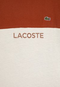 Bavlněné tričko s rezavou horní částí a krémovou dolní částí. Obsahuje logo Lacoste a má elegantní, hladkou texturu.