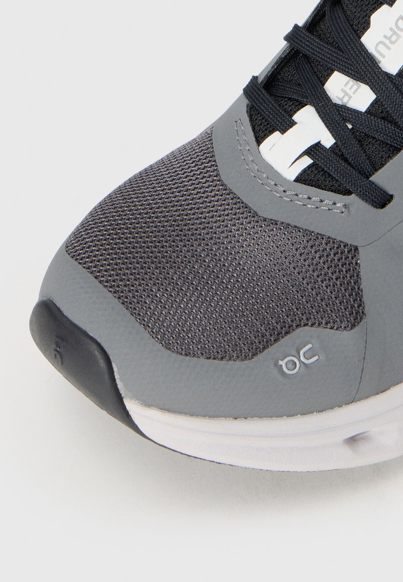 Grau sportlicher Schuh mit einem strukturierten Mesh-Obermaterial, glatten synthetischen Akzenten, schwarzen Schnürsenkeln und einer weißen Sohle mit Logodetail an der Seite.