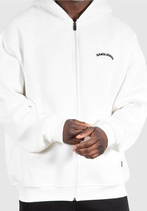 Personne portant un sweat à capuche blanc zippé avec le logo « SMILODOX » sur la poitrine, les mains légèrement jointes devant, sur un fond uni.