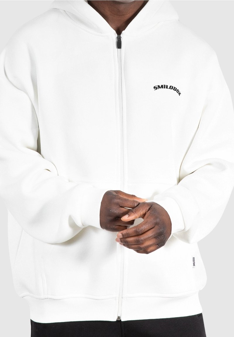 Personne portant un sweat à capuche blanc zippé avec le logo « SMILODOX » sur la poitrine, les mains légèrement jointes devant, sur un fond uni.