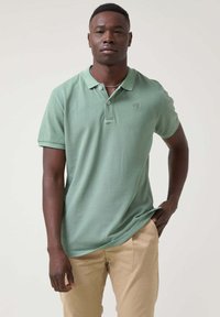 Camisa polo de algodón verde con mangas cortas, tapeta de tres botones y detalle del logo en el pecho, combinada con pantalones beige claros.
