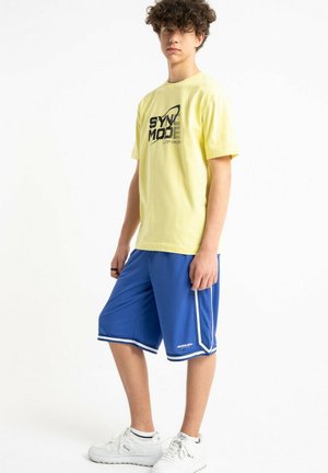 Młody człowiek z kręconymi włosami, ubrany w żółtą koszulkę z nadrukiem, niebieskie spodenki do koszykówki z białymi paskami oraz białe trampki, stojący na białym tle.