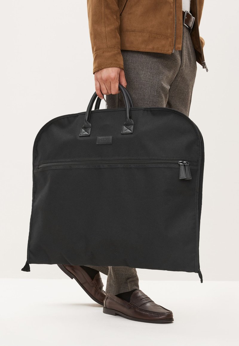 Personne portant une veste et un pantalon marron tenant un grand sac à vêtements noir zippé par les poignées sur le côté.