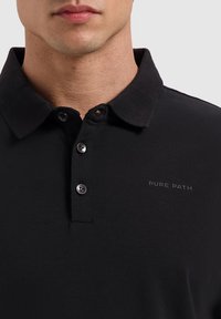Zwart poloshirt met een klassieke kraag, drie-knopen sluiting en een subtiele "PURE PATH" logo op de borst. Gemaakt van zachte stof.