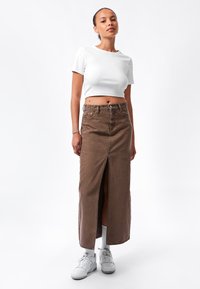 Falda maxi de denim marrón con abertura frontal, combinada con un top corto blanco. La modelo lleva zapatillas blancas y medias hasta la rodilla. Textura suave y estilo casual.