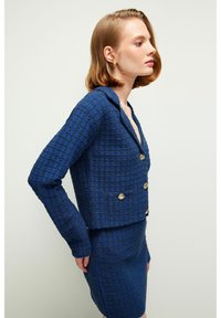 Blazer de tweed azul con estampado y botones dorados, con cuello con muesca y un diseño recortado, acompañado de una falda ajustada a juego.