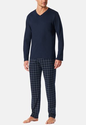 Uomo che indossa una maglia a maniche lunghe blu navy con scollo a V e pantaloni blu navy con motivo a griglia bianca, in piedi a piedi nudi su uno sfondo semplice.