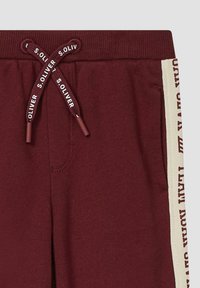 Bordeauxrote Jogginghose mit gerippetem Bund, ausgestattet mit einem Kordelzug mit Markenname. Seitliche Streifen in Creme mit braunem Textmusterakzent.