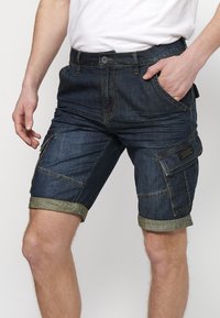 Homme portant un short cargo en denim bleu foncé avec des revers roulés, debout avec une main dans la poche et t-shirt blanc partiellement visible.