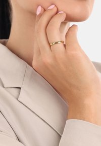 Primo piano di una mano con unghie rosa chiaro che indossa un semplice anello d'oro, toccando il mento, con un blazer beige visibile.