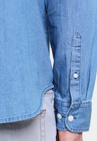 Camicetta in denim azzurro chiaro con maniche lunghe, dotata di bottoni bianchi e orlo curvo. La trama è liscia, con una vestibilità casual.