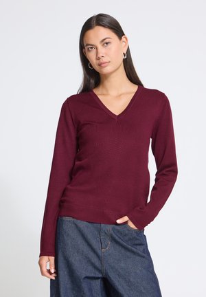 Jeune femme portant un pull bordeaux à manches longues et col en V ainsi qu'un jean bleu décontracté, debout avec une main dans la poche sur un fond uni.